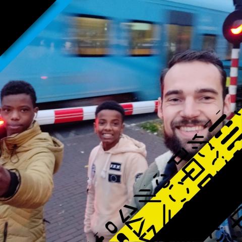 afbeelding van een man en twee jongens bij een spoorwegovergang met daarachter een blauwe trein
