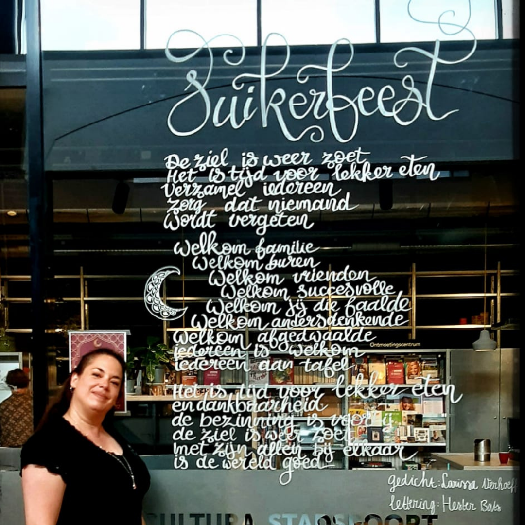 foto van een vrouw met een zwart t-shirt en lang donker haar. Ze lacht en staat voor een raam waarin in handlettering het gedicht Suikerfeest staat van Larissa Verhoeff.